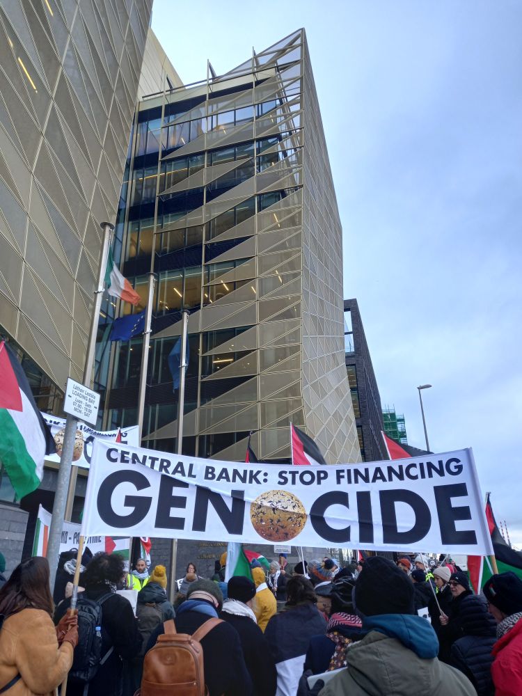 Dublin, outside Central Bank building entrance (tall fancy glass building) protesters with Palestinian flags and banner CENTRAL BANK: STOP FINANCING GENOCIDE 