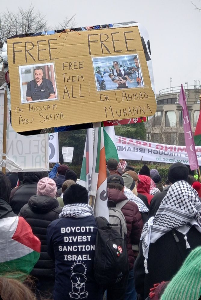 Dublin, Ireland 
In front of US Embassy, crowd gathering with anti-genocide banners 
and Palestinian flags, in the forefront a protester wearing a jacket with BOYCOTT DIVEST SANCTION ISRAEL at the back. 
Insert with a cardboard sign with pictures of Dr Huassam Abu Safiya and Dr Ahmad Muhanna and written FREE FREE FREE THEM ALL
