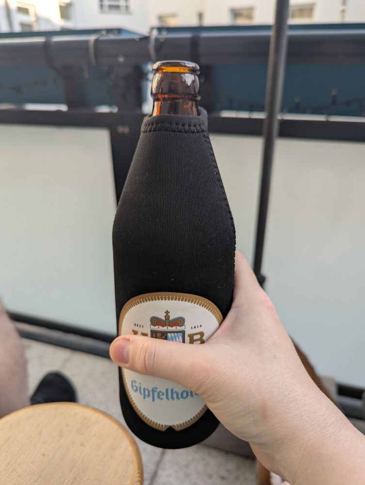 A 500ml bottle of Augustiner Helles in a Hofbräuhaus Traunstein cozzie Held in my hand as I sit on the balcony enjoying the Friday evening.