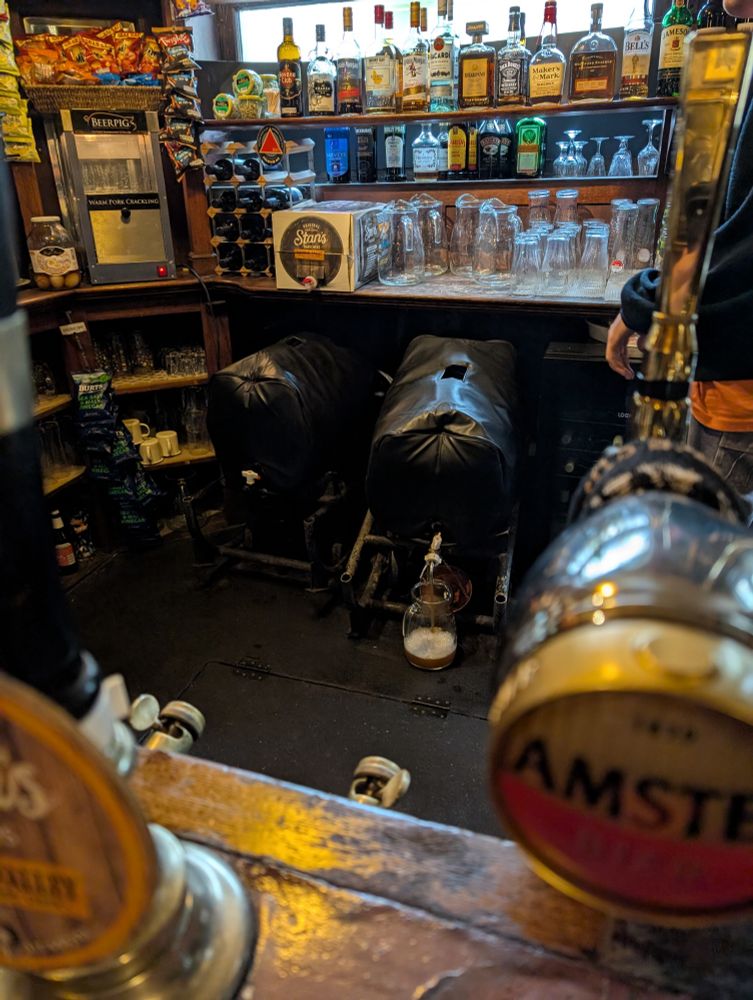 Two Bass casks with cooling jackets on and a partially filled glass jug in front of one. The Bass is gravity poured from the cask.