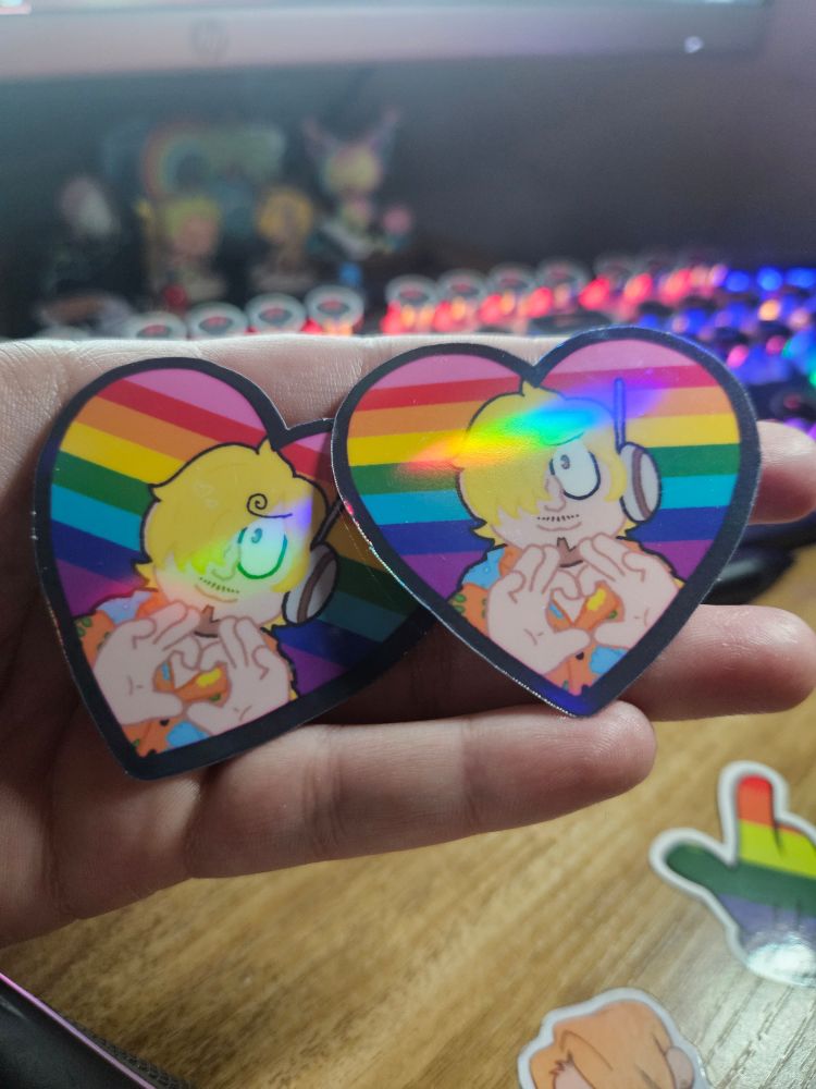 photo of me holding two heart shaped stickers of sanji in his egghead outfit with a gay pride flag background 