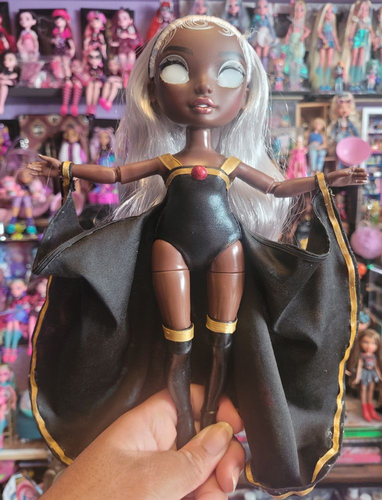 Rainbow high Ayesha Sterling Doll turned into Custom X-Men Storm. Wearing a black leotard with black thigh highs and cape, trimmed with gold ribbon. Doll shown floating in person's hand.