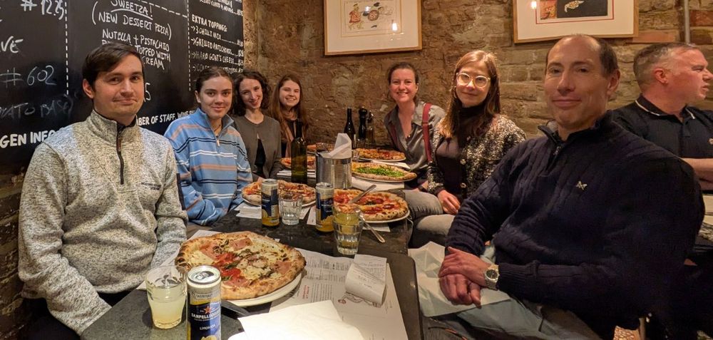 A group of eight people is seated around a table in a restaurant with exposed brick walls. They are enjoying a meal together, with several large pizzas, drinks, and menus spread across the table. The group consists of men and women dressed casually, smiling at the camera. A chalkboard menu with handwritten items is visible in the background, along with framed artwork on the walls. 