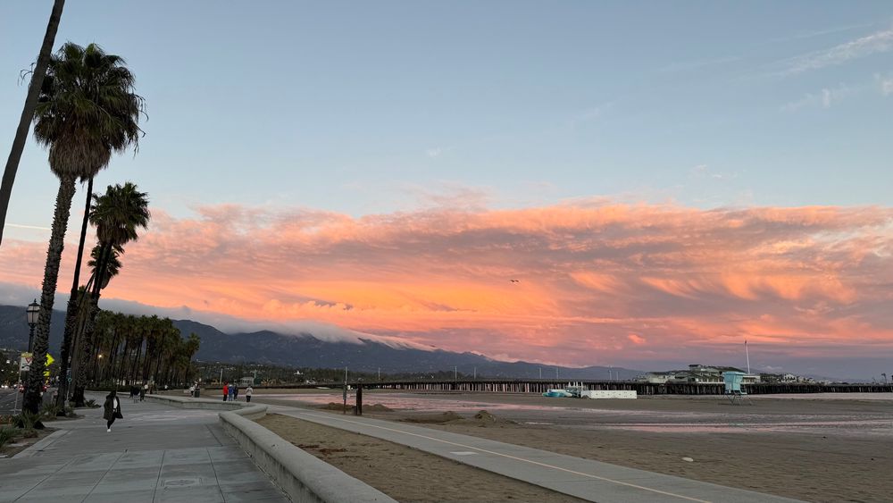 Photo of Santa Barbara’s West beach shoreline with big pinkish orange clouds and Sterns Wharf and beach in foreground. 