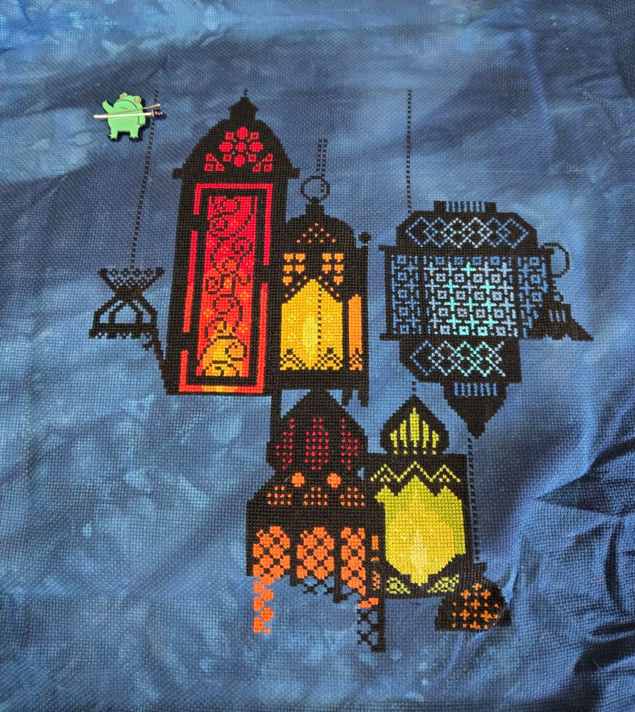 Moroccan style lantern outlines are cross stitched in black with various colors inside the lanterns including reds, oranges, blues and greens. There is a magnetic needle minder in the top left corner, shaped like a frog giving the bird, with a needle and a decorative pin attached to it.