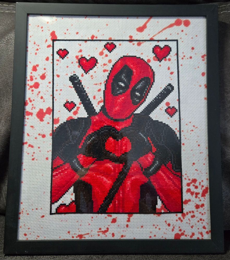 A cross stitch picture of Deadpool making a heart with his hands with hearts floating around him, made on white fabric that looks blood-spattered