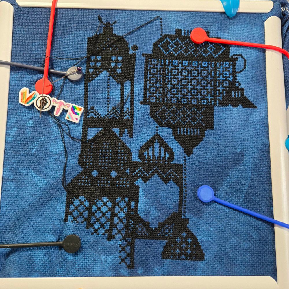 A partially completed cross stitch of Moroccan style lanterns - black thread on tie-dyed blue aida material