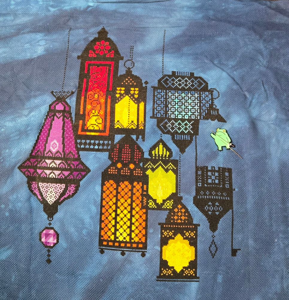 Moroccan style lantern outlines are cross stitched in black on blue tie-dyed fabric, with various colors inside the lanterns including purples, reds, oranges, blues and greens. There is a magnetic needle minder in the middle right of the design, shaped like a frog giving the bird, with a needle and a decorative pin attached to it.