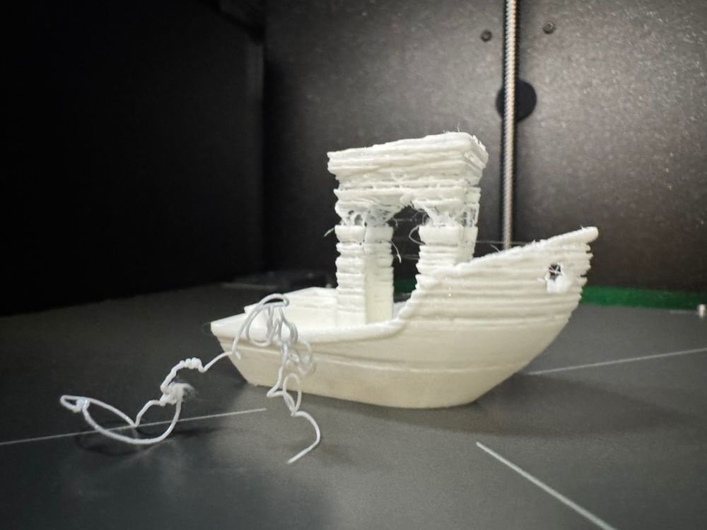 a picture of a white, freshly 3d printed 3dbenchy, still on its build plate. it is clearly under-extruded; the higher up you go on the print, the more material is missing. the chimney is missing entirely, and you can see a string of filament in front of the benchy, presumably what was supposed to be the chimney.