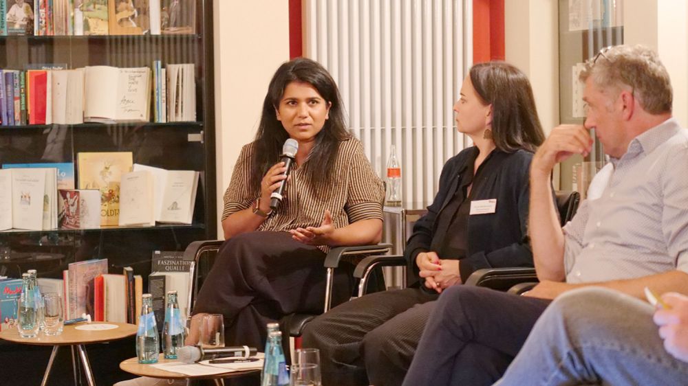 Vor einer Bücherwand sitzt eine Frau neben Menschen auf dem Podium und spricht in ein Mikrofon.
