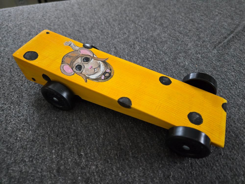 Wedge shaped wooden car painted to look like a block of yellow cheese. A painted mouse as a driver wearing goggles and hat.