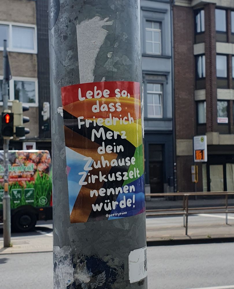 Sticker an einem Laternenpfahl: Lebe so, dass Friedrich Merz dein Zuhause Zirkuszelt nennen würde! @poetryrune  in weißer Schrift auf buntem Hintergrund, angelehnt an die Pride Flagge. Hinter dem Laternenpfahl ist eine mehrspurige Straße mit Fußgängerampel und Bushaltestelle zu sehen, dahinter eine Häuserfront.