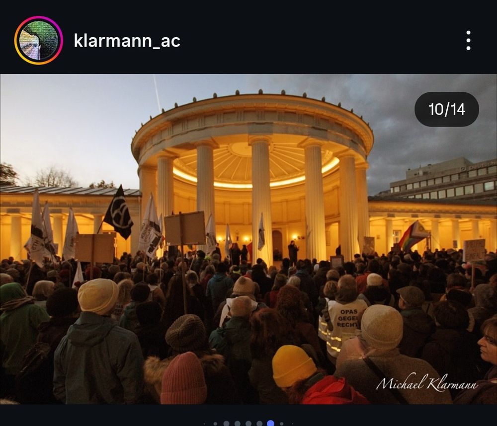 Screenshot eines Insta Beitrags von klarmann_ac  Viele Menschen bei einer Kundgebung. Es dämmert. Im Hintergrund ist deutlich die Rotunde des Elisenbrunnens zu sehen, sie ist im Innern mit angenehm warmem Licht angestrahlt. Signatur unten rechts: Michael Klarmann