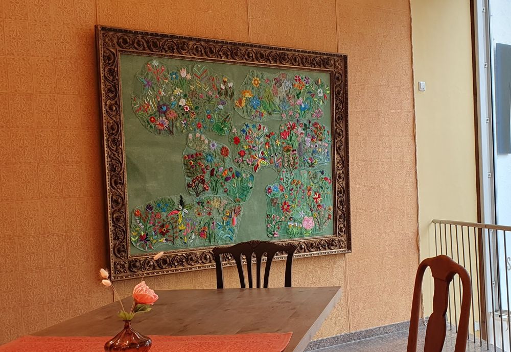 Zwei Stühle und ein Tisch im Vordergrund. Im Hintergrund an der Wand ein gerahmtes Kunstwerk: viele bunte filigran gestickte Blumen auf grünem Hintergrund. Auf dem Tisch steht eine Blumenvase. Die Wand ist in gelb-orangem Farbton tapeziert.