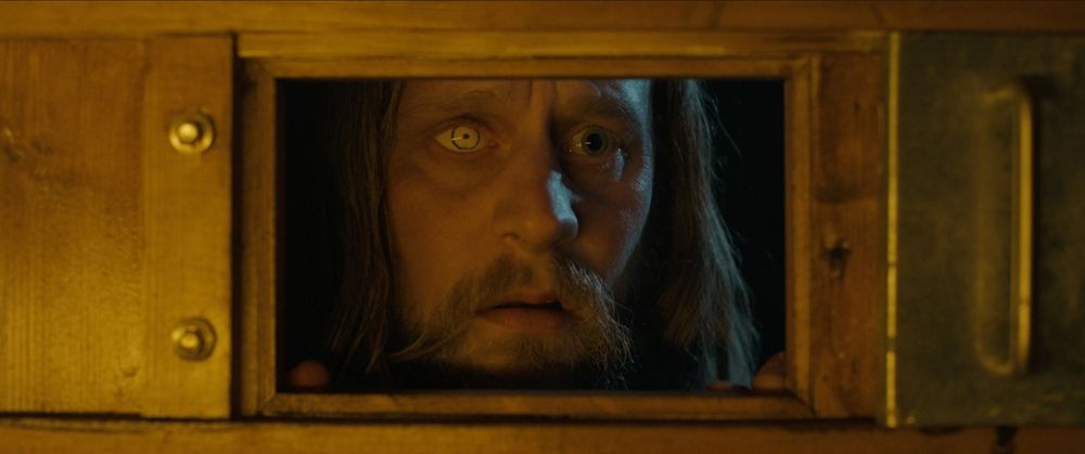 A man peers through a hatch in a door. He has a glass eye, long hair, and a beard. He looks anxious