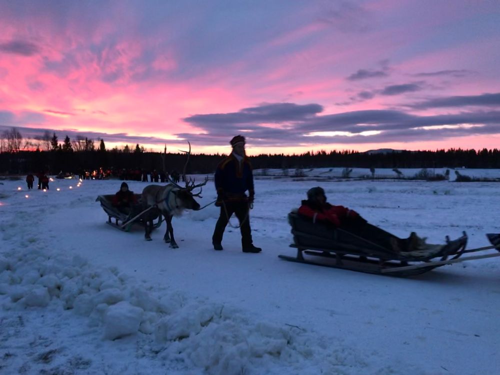 A serene winter scene in Lapland at sunset, featuring a person leading a reindeer pulling a sled with another person sitting inside. The sky is painted with vibrant hues of pink and purple, creating a picturesque backdrop. Snow covers the ground, and in the distance, more people and lights suggest a gathering or event, highlighting the natural beauty and traditional culture of Lapland.