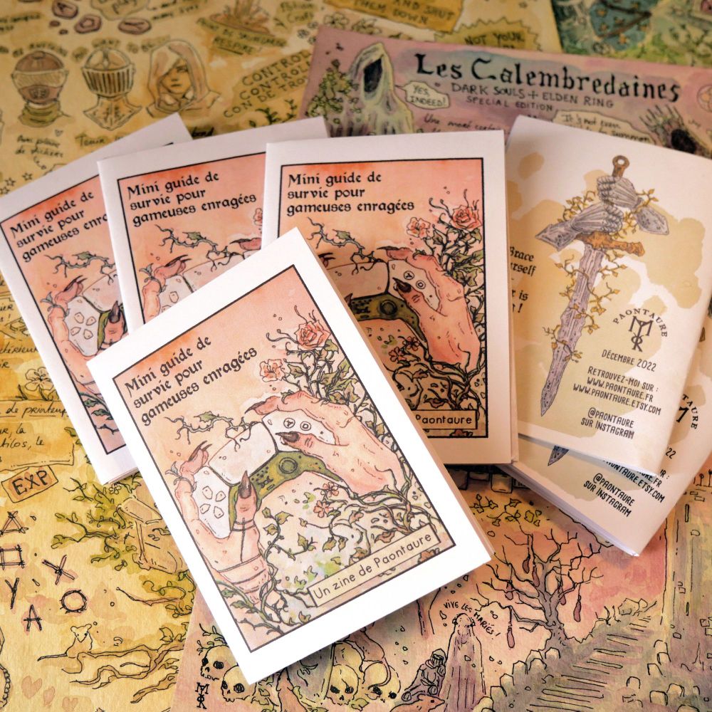 Photo d'un tas de mini-zines "Mini guide de survie pour gameuse enragées" en vrac sur des planches de croquis aquarellisés. La couverture des zines montre une manette de PS5 entre des mains de sorcière.