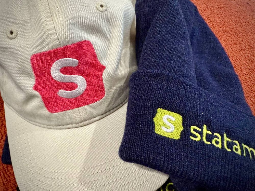 A cream baseball cap and a navy blue knitted cap, both with the Statamic logo on them