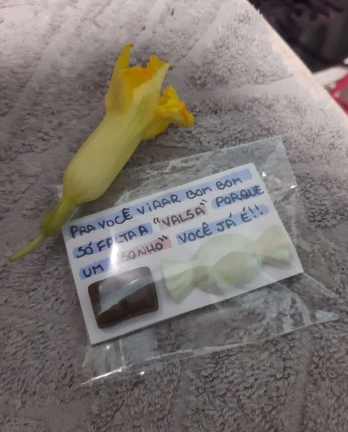 Foto de uma florzinha amarela e um cartãozinho com uma cantada brega escrito"pra você virar bom bom só falta a 'valsa' porque um 'sonho' você já é" e tem um mini chocolate e um mini bom bom de biscuit