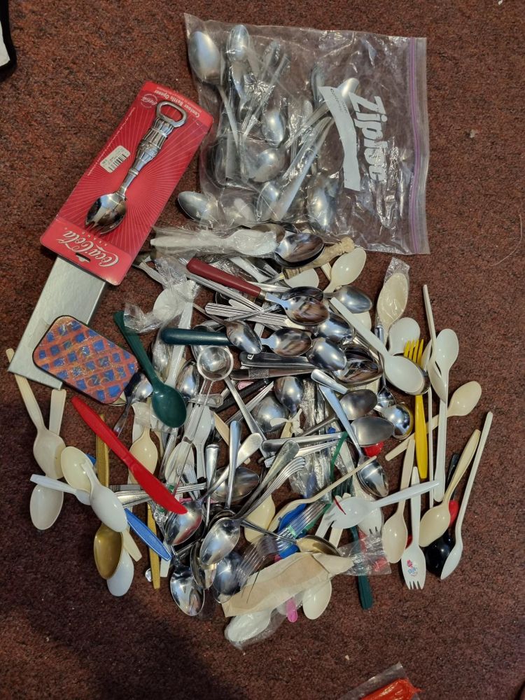 A photo of a huge pile of spoons on a carpeted floor. There are spoons of many sizes, materials, colors, and shapes. So many spoons.