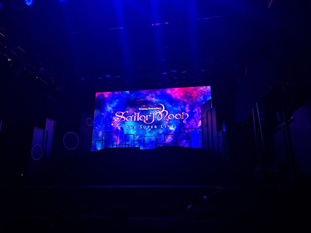 Photo of a stage with a glowing screen showing the Pretty Guardian Sailor Moon The Super Live title card against a multicolor cosmic background. There are black outlines of a city skyline along the bottom 