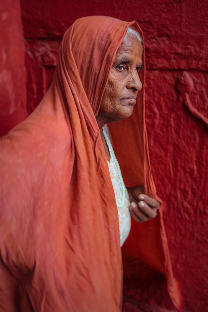 Elderly woman in a red-orange sari walking past a textured red wall in India, captured in profile
