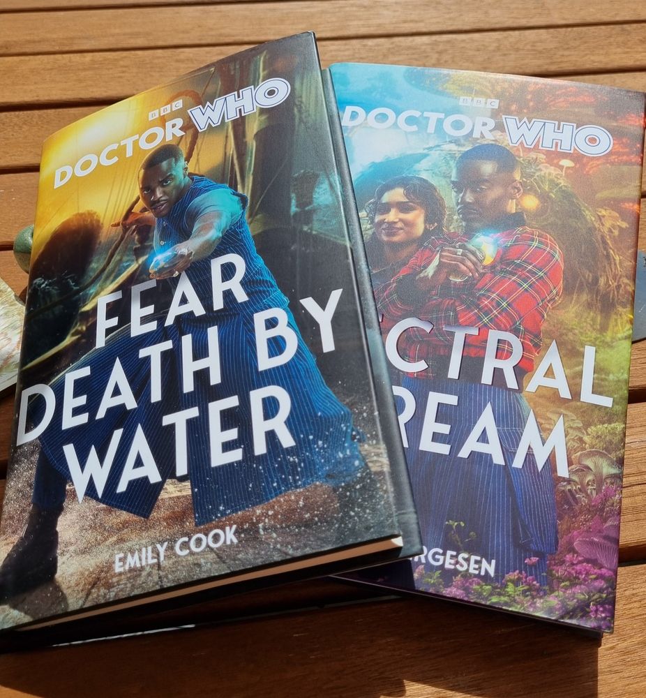 Wooden garden table with two Doctor Who books on. First is Fear Death By Water by Emily Cook and the second is Spectral Scream by Hannah Fergesen. Both feature the Fifteenth Doctor and Belinda.