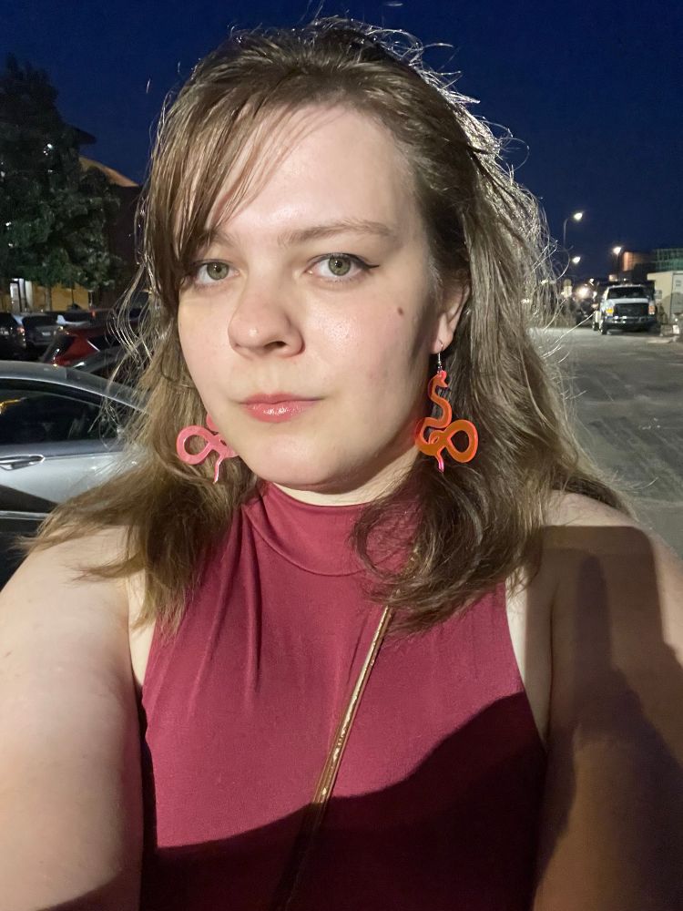 charlotte on the sidewalk at night wearing a red sleeveless top and translucent pink earrings shaped like snakes