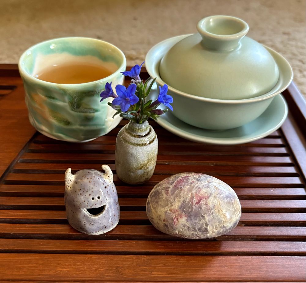 Gong fu tea set on a dark brown bamboo tea tray, including a green gaiwan, a green and white tea cup full of golden colored tea, a small bouquet of blue flowers, a unicorn pegmatite stone…and the main attraction: the happiest tea pet ever! He is a little monster guy, with small white horns, a round purplish grey glazed body, and the goofiest grin.
