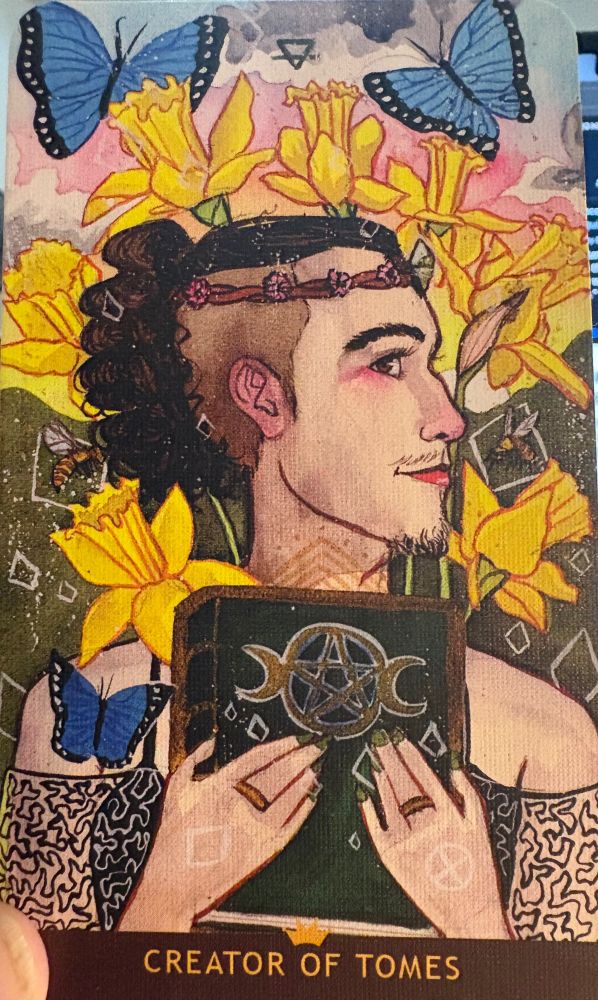 The Creator of Tomes from the Numinous Tarot deck. A white genderqueer person is holding up a green book with a golden pentacle and 2 crescent moons on its cover. Their black hair is long and pulled back to reveal their shaved undercut. They are wearing a crown with tiny blossoms on it. They have a mysterious, magnanimous smile on their face, with a light mustache and careful makeup. They are wearing a spaghetti strap tank with a black lace shawl around their shoulders. Behind them are bright yellow daffodils and large blue butterflies.