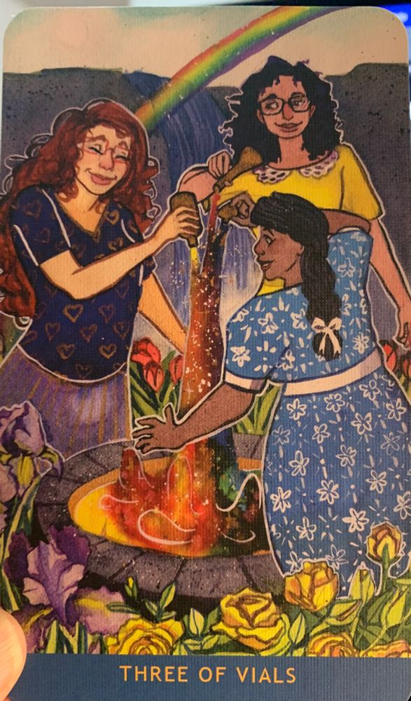 The Three of Vials from the Numinous Tarot deck. It shows three people surrounding a cauldron, each of them pouring a vial into the swirling rainbow liquid. The three people include a red-haired white femme wearing a blue shirt covered in hearts, a brown woman wearing a light blue dress covered over in a white floral pattern, and a third figure wearing yellow, with curly hair to their shoulders. A rainbow arcs over all of them as they mix the ingredients of their vials together.