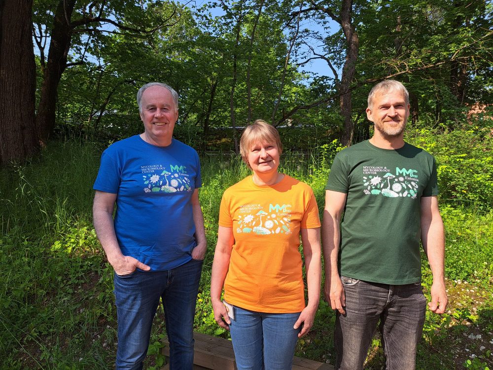 Image of three people wearing a blue, an orange and a green T-shirt. The design on the shirts is the same by featuring the logo for MMC and some fungi and tiny creatures, e.g. bacteria on the chest.