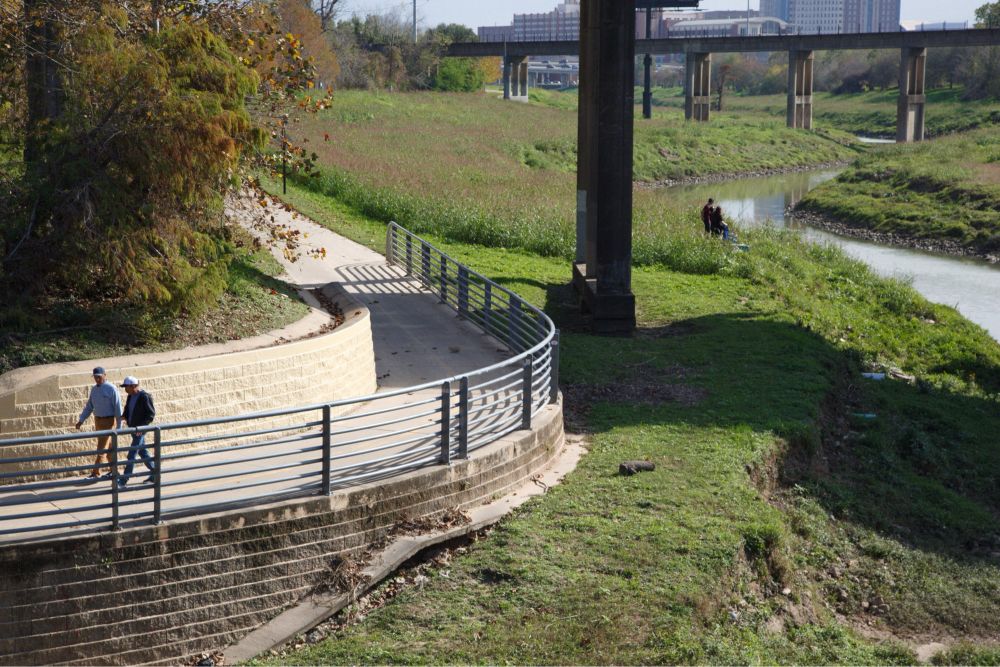 A riverside pedestrian walkway at White Oak Bayou in Houston, Texas, where two people are strolling along, bordered by metal railings and grassy slopes, with a concrete bridge and urban buildings visible in the distance.