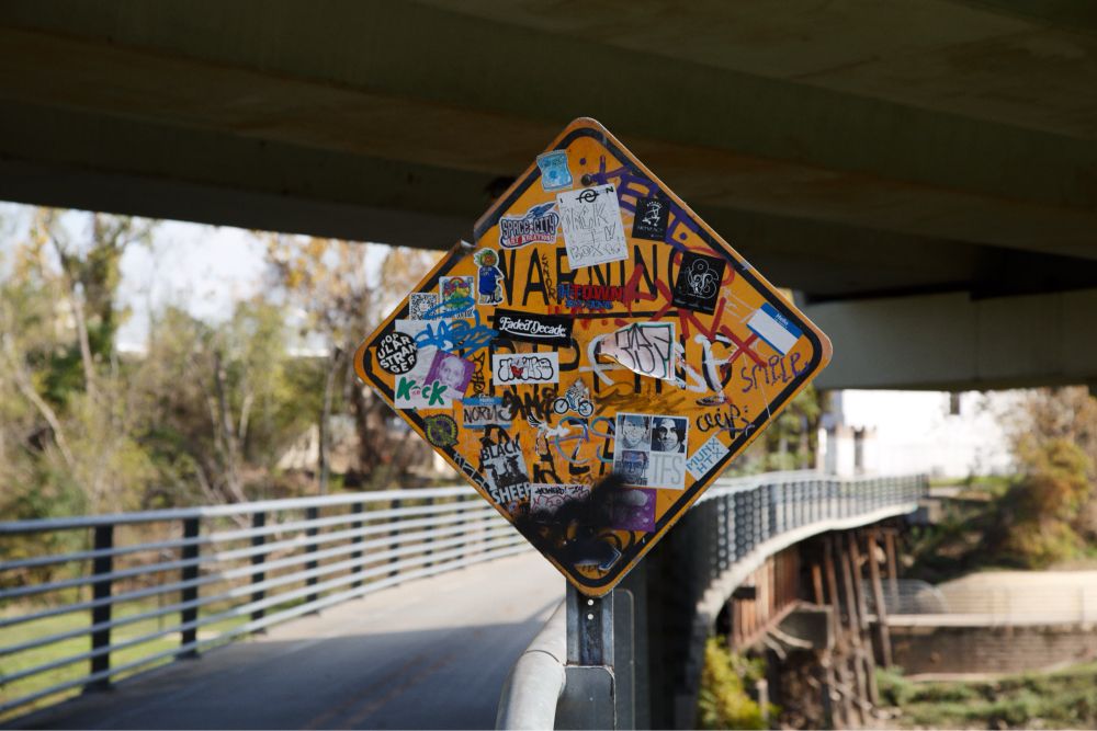 A yellow diamond-shaped road sign covered with colorful stickers and graffiti, placed near a bridge and walkway at White Oak Bayou in Houston, Texas, with greenery and railings in the background.