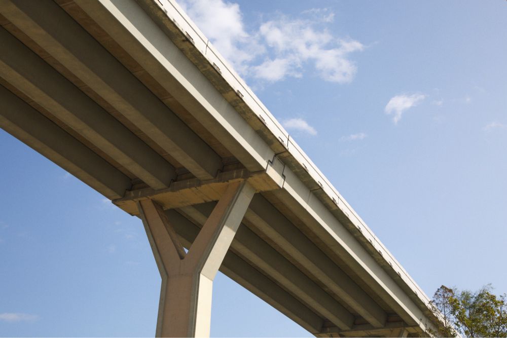 A view from underneath a large concrete highway overpass near White Oak Bayou in Houston, Texas, showing its thick beams and sturdy Y-shaped support structure against a bright blue sky.