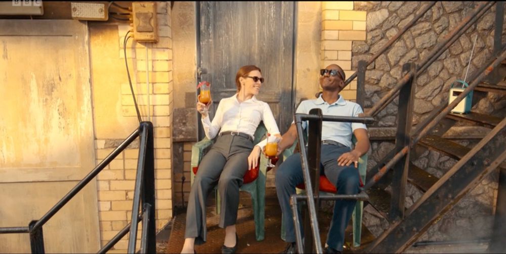 Screen capture showing the Doctor (Ncuti Gatwa) enjoying drinks with Anita (Steph de Whalley) on a fire escape, presumably at the Sandringham Hotel.