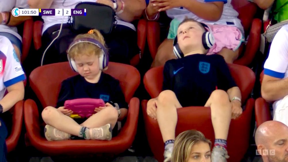 Screen capture of two children sitting in plush reddish chairs at the football stadium. Both wear dark England shirts and over-the-ear headphones. The one of the left is enraptured with his pink-encased tablet computer. The one on the right has tipped his chair seat up and is looking up at the sky.