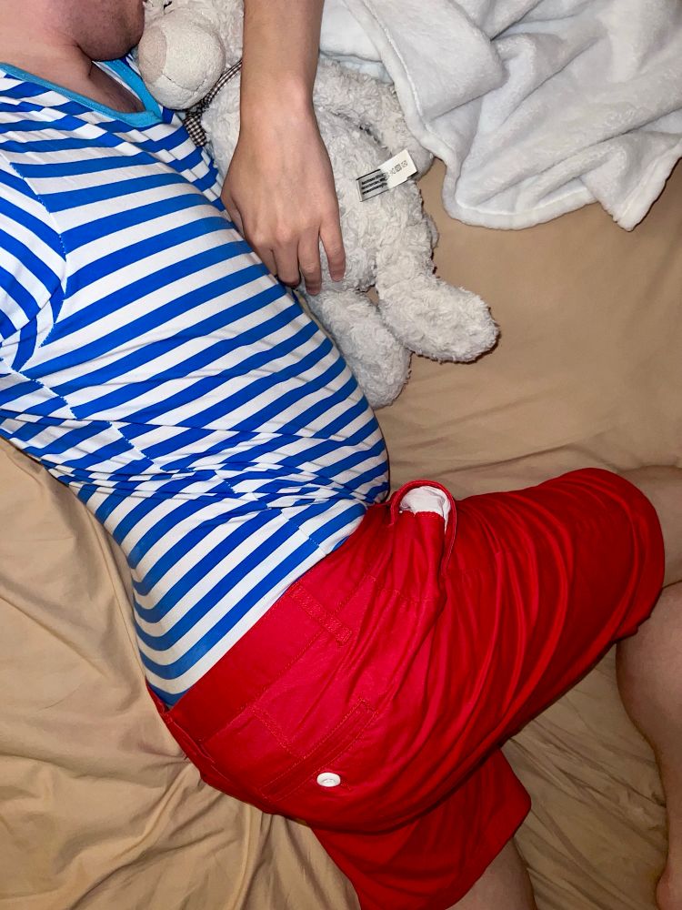 Me laying on my side in bed in my blue and white striped onesie and red shorts, with a soggy Teddy’s Ultra nappy underneath, cuddling Teddy and my blanket