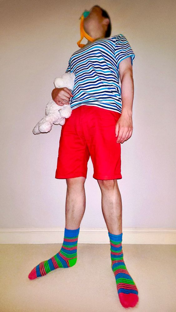 Me stood in a blue and white striped t-shirt, red shorts, and blue, green and pink striped socks, with a soggy Animooz nappy underneath, sucking my turquoise dummy and holding Teddy