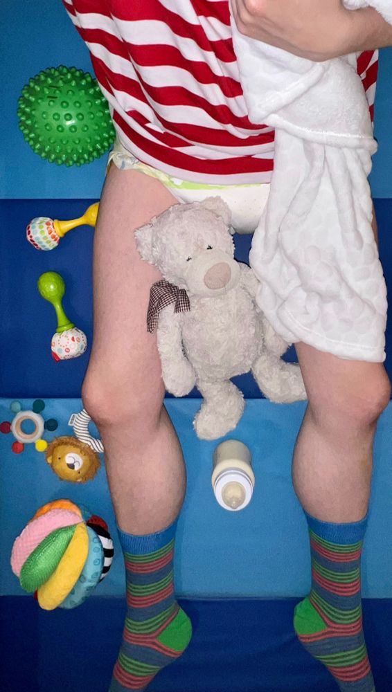 Me sat on the floor in a red and white striped t-shirt, Animooz nappy, and blue, green and pink striped socks, holding my blanket with Teddy, my bottle and some toys
