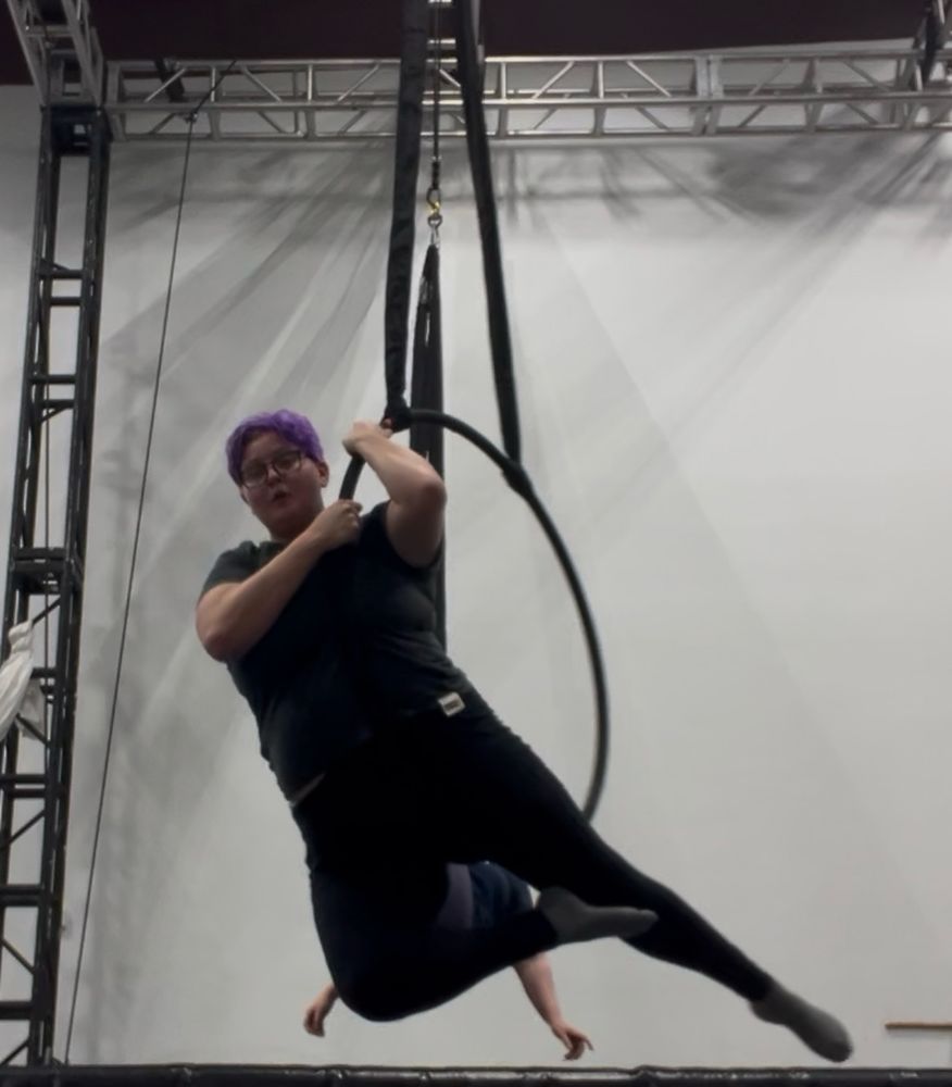 Mayson, a purple haired enby with black square glasses, holds themself up in a Lyra facing the camera with both arms while their legs are pointed in an arrow shape
