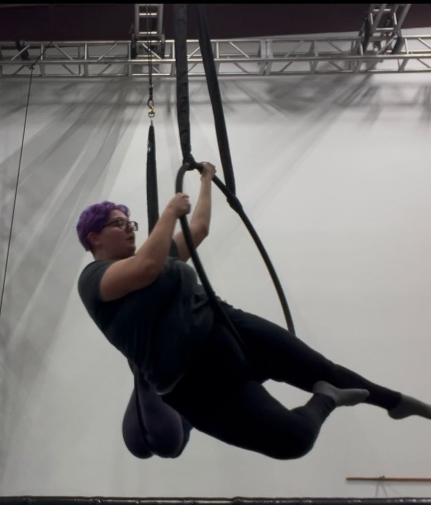 Mayson, a purple haired enby with black square glasses, holds themself up on the left side of a Lyra with both arms while their legs are pointed in an arrow shape
