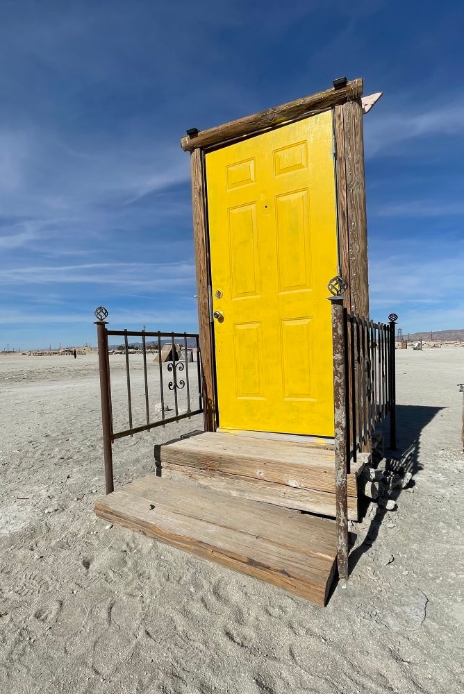 This image is of the artwork “Open House” a yellow door inside a door frame with wooden steps and railings unattached from a house with a bright blue sky and desert scenery behind it 