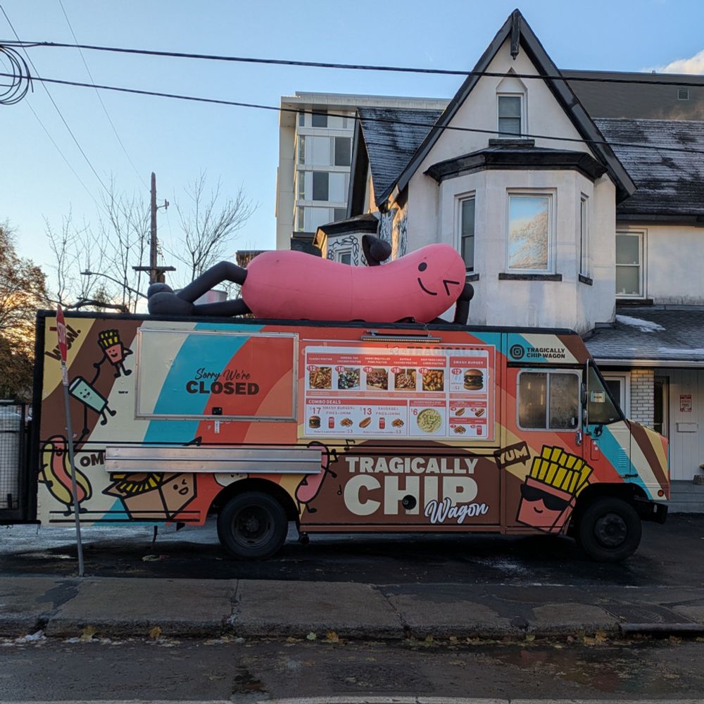 Rainbow-coloured food truck called "Tragically Chip Wagon." A giant inflatable hot dog reclines on top, winking enigmatically.