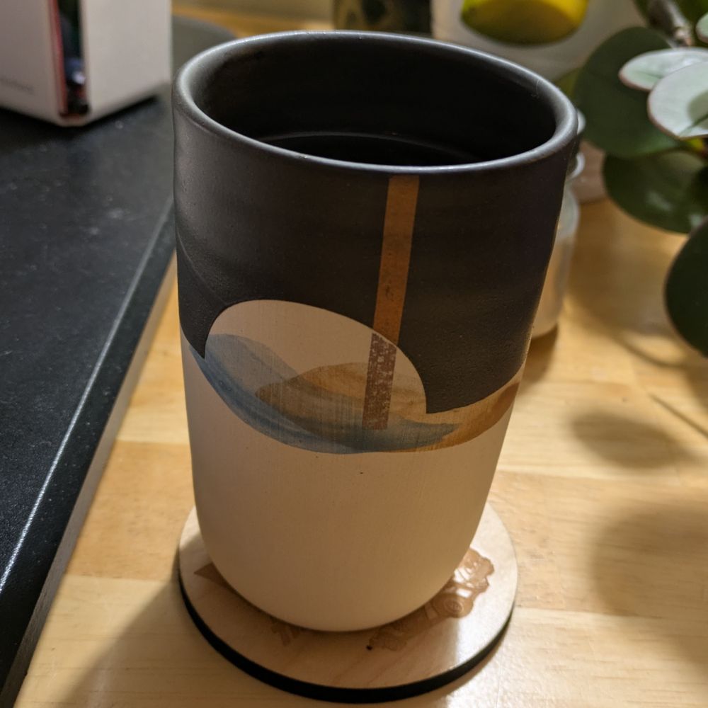 Pottery coffee mug, painted black on top with a watercolour-style image of mountains and sun. It sits on a wooden Back to the Future coaster.