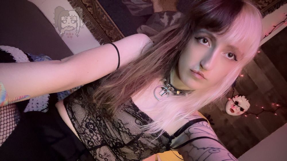 Sleepy with her split-dyed hair down staring into the camera. She is wearing a lacy, black strappy top and a spiked, black collar. You know she has fishnets on.