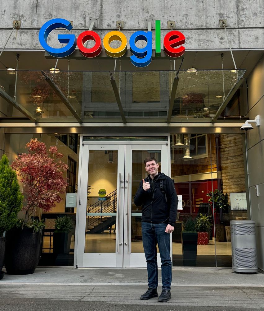 A man stands smiling outside a Google office, giving a thumbs-up. He wears a black jacket and jeans, with the colorful Google logo above the glass entrance. Potted plants frame the doorway.
