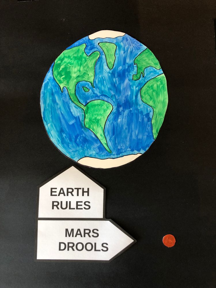 Hand-painted sign with earth on a black background, with a small reddish planet off to the side. Text pointing at Earth reads, Earth Rules.” Arrow pointing at reddish planet with text says, “Mars Drools.”