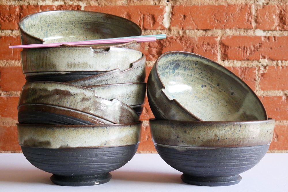 A tall stack of 6 ramen bowls on the left, each with black clay exposed on the outside, a deep angular foot ring and carved texture - then an organic white/brown glaze with blue specks around the notched rim and inside. the stack is aesthetically off-kilter, and the top bowl has chopsticks laid across two notches in the rim. on the right, just two bowls of the same design, stacked a you can see more of the glazed inside. 