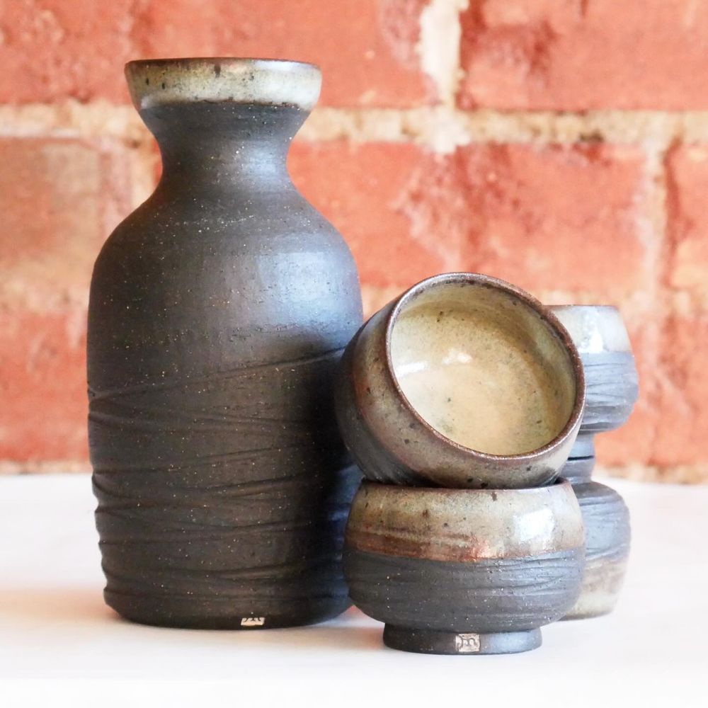 Black clay sake set, a small mostly bare bottle with rounded shoulders into a neck and then out to a cup-like lip, glazed with an organic white that breaks brown at the rim. Beside it, four small cups stacked in pairs, a narrow foot holds a small bowl shape - again bare with an icy white/brown rim and inside